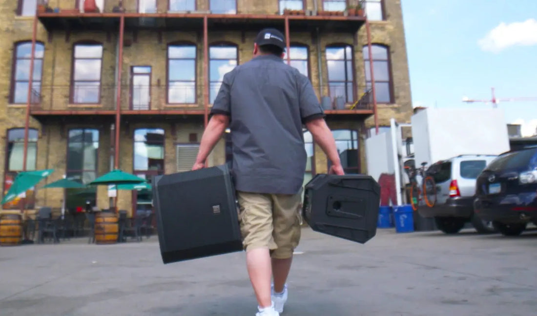 Person carrying two black cases in an urban setting with a building and cars in the background.